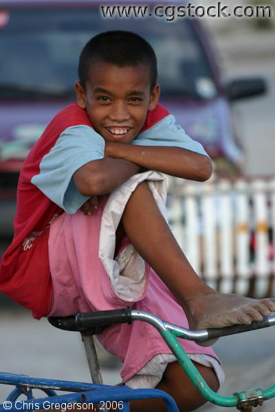 Image of a Smiling Street Boy on a Bicycle in Front of a Red Car with Dirty Feet